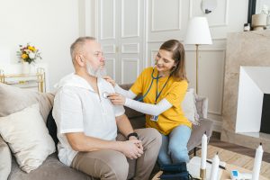 A healthcare worker in a yellow shirt is using a stethoscope on an elderly man sitting on a sofa. The room is bright and decorated with flowers.