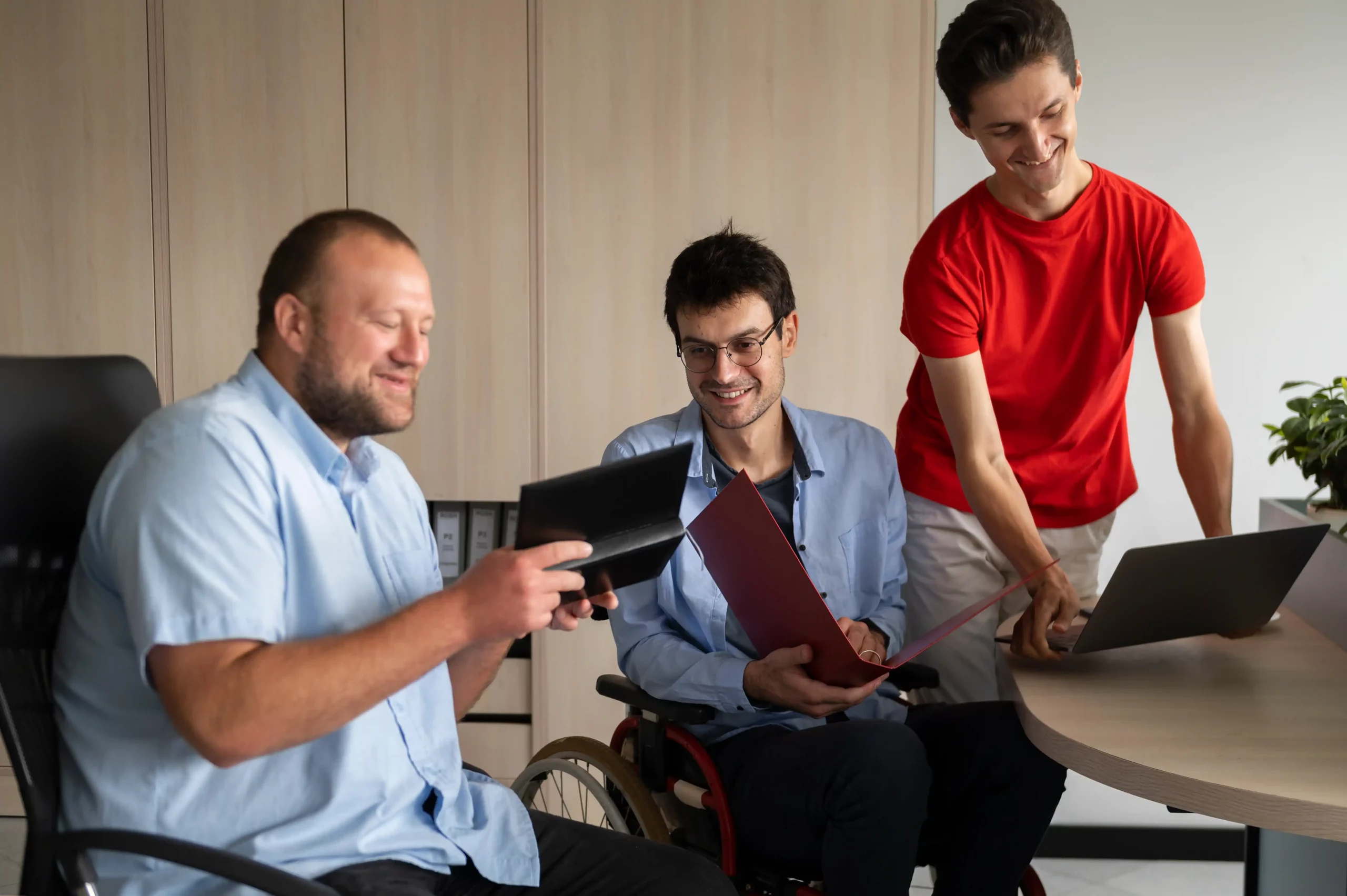 Three men in wheelchairs are seated at a table, each using a laptop for work or collaboration.