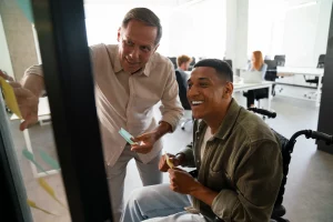 Two colleagues, one in a wheelchair, collaborate by placing sticky notes on a glass wall in a bright, modern office, conveying teamwork and inclusivity.