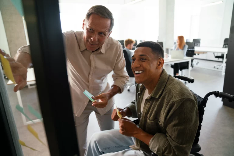 Two colleagues, one in a wheelchair, collaborate by placing sticky notes on a glass wall in a bright, modern office, conveying teamwork and inclusivity.