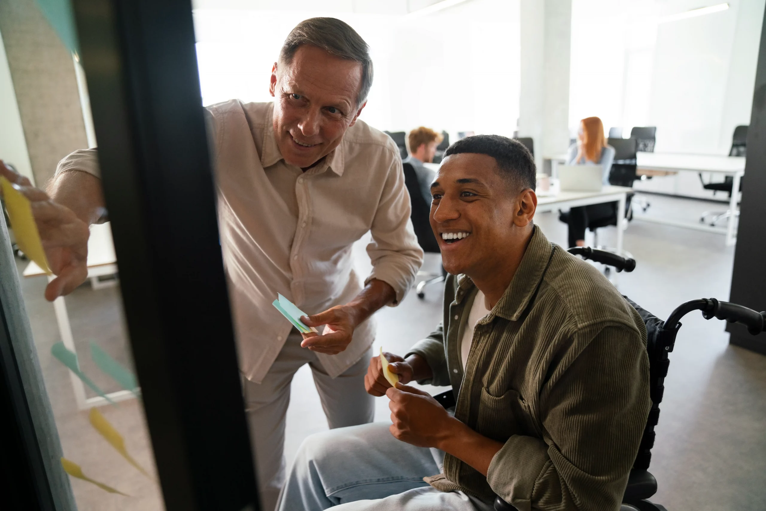 Two colleagues, one in a wheelchair, collaborate by placing sticky notes on a glass wall in a bright, modern office, conveying teamwork and inclusivity.