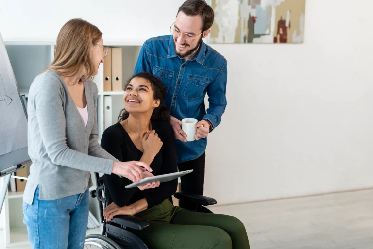 Two women stand beside a man in a wheelchair, all three smiling and interacting in a friendly manner.
