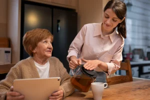 An elderly woman with a tablet smiles at a younger woman pouring tea into a mug. The scene conveys warmth and companionship in a cozy kitchen.