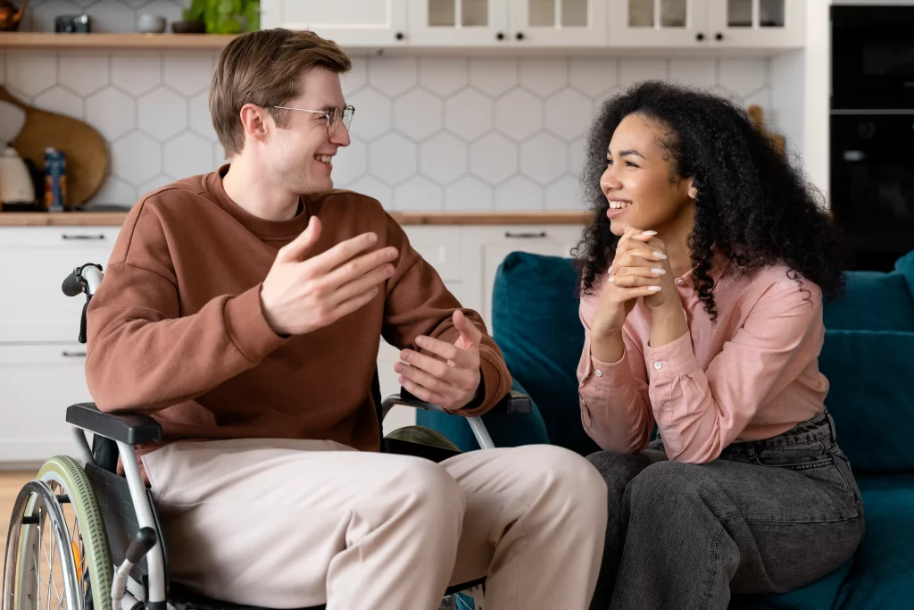 A man in a wheelchair and a woman sit in a modern kitchen, engaged in a lively conversation. Both are smiling, conveying a warm and friendly atmosphere.