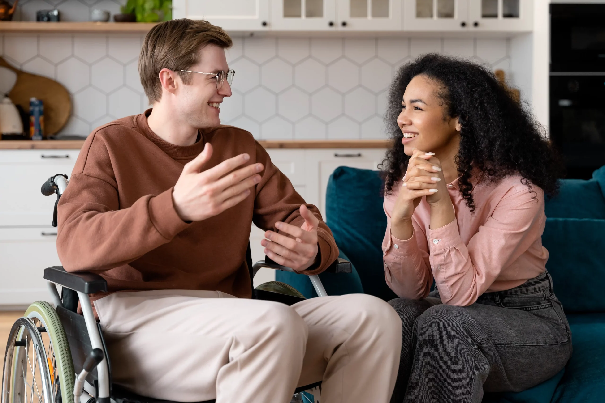 A man in a wheelchair and a woman sit in a modern kitchen, engaged in a lively conversation. Both are smiling, conveying a warm and friendly atmosphere.