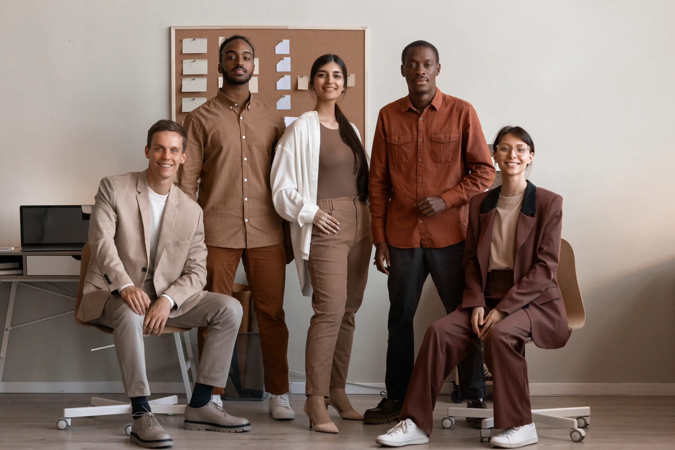 A group of five diverse people in business casual attire pose confidently in an office with neutral tones. They're smiling, exuding teamwork and positivity.