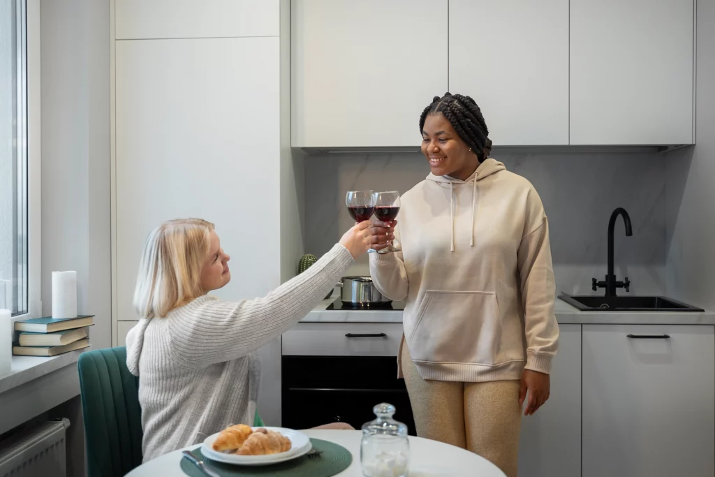 Two people in a modern kitchen toast with red wine glasses, smiling. One sits with croissants on a plate; the other stands in a hoodie. Cozy and cheerful.