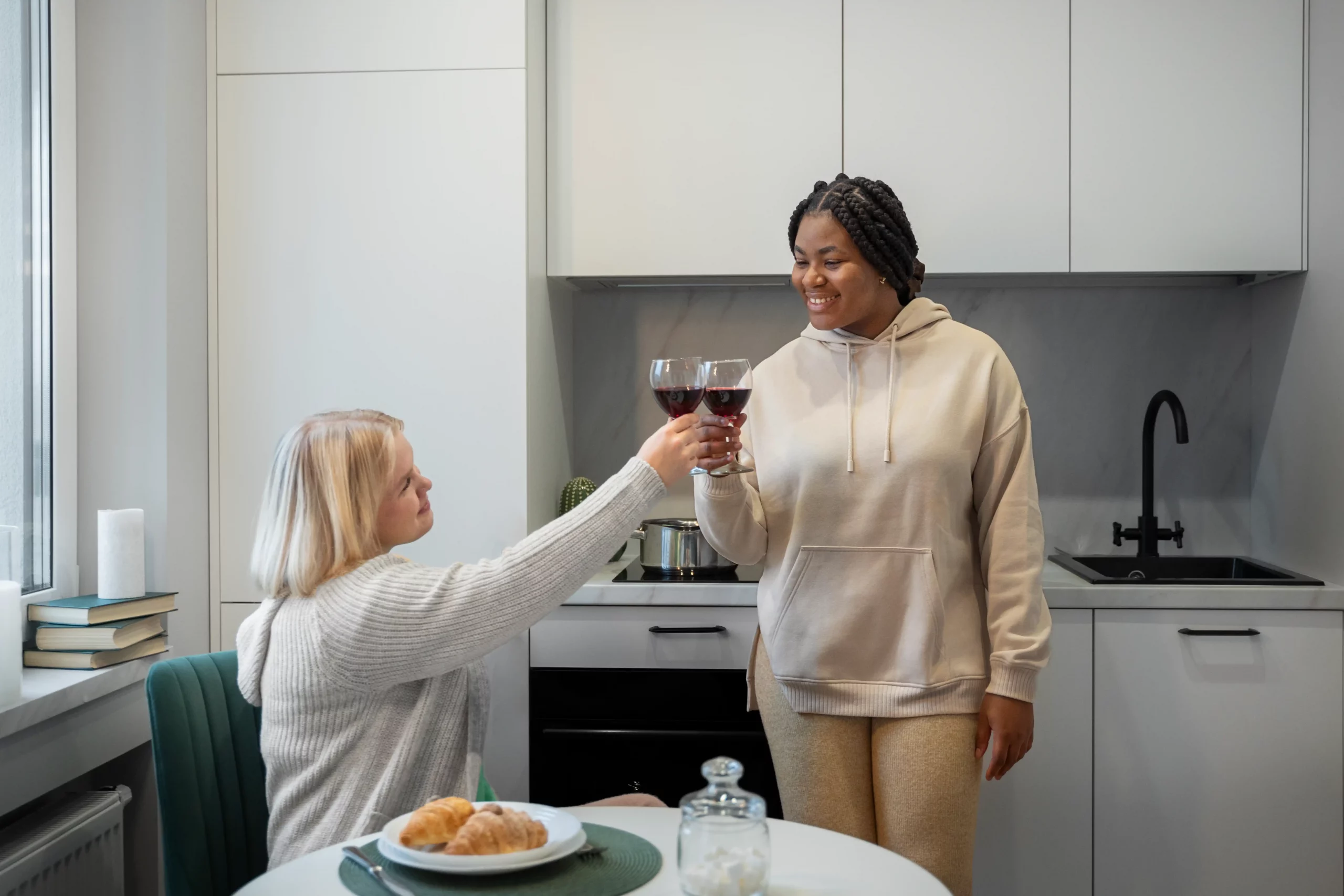 Two people in a modern kitchen toast with red wine glasses, smiling. One sits with croissants on a plate; the other stands in a hoodie. Cozy and cheerful.