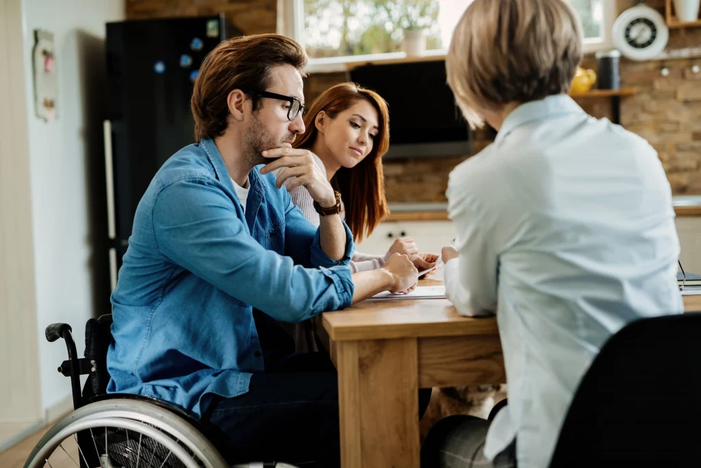 A man in a wheelchair and a woman sit across from another person at a table, engaged in discussion. The mood is thoughtful and collaborative.